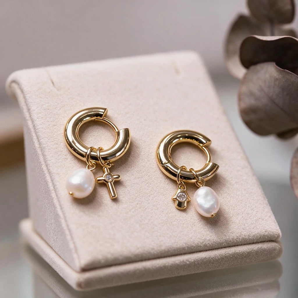 Mismatched gold-tone charm hoop earrings with cultured baroque pearls, a cross charm and a hamsa charm, on a cream jewellery stand