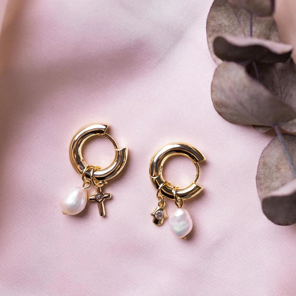 Pair of gold-tone charm hoop earrings on pink silk, showing the cross and hamsa charms alongside baroque pearl drops